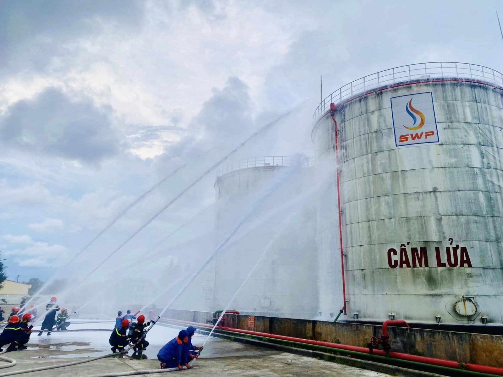 Các tia nước và bọt trắng xóa bao phủ khu bồn chứa xăng dầu mang logo S.W.P, thể hiện khả năng ứng phó hiệu quả với tình huống cháy nổ quy mô lớn.