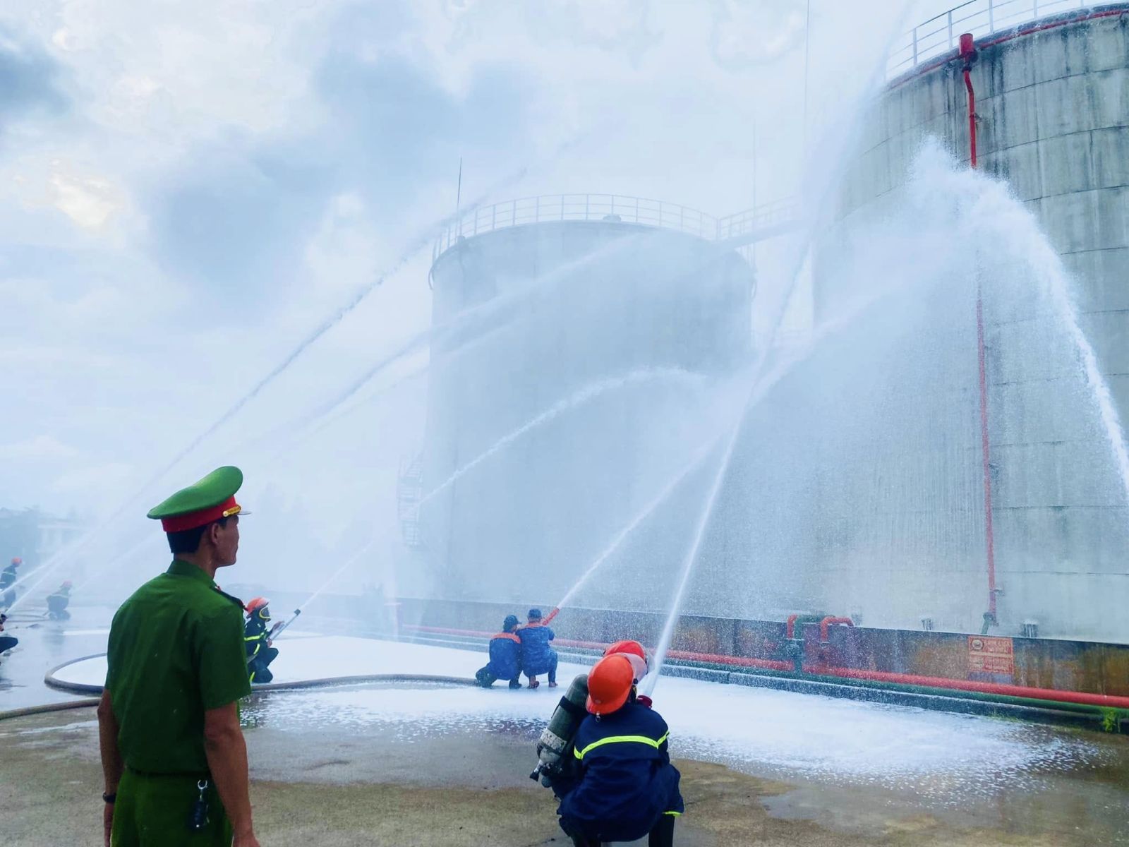 Lực lượng PCCC đồng loạt triển khai vòi nước và bọt chữa cháy hướng về khu vực bồn chứa, mô phỏng tình huống cháy lớn.
